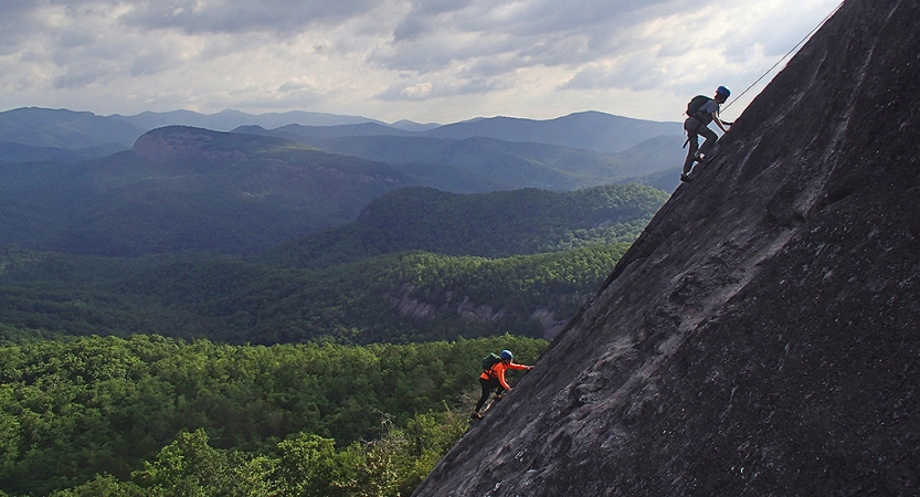 Two people wearing safety gear ascend up a step rock slope with the blue ridge mountains below them. 